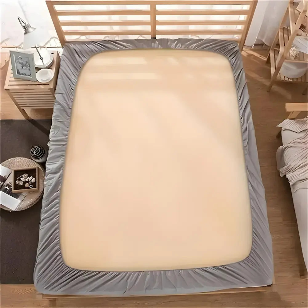 Imagem de uma cama vista de cima, com um colchão coberto por uma capa cinza clara nas bordas e uma parte central bege. A cama tem cabeceira de madeira e está em um quarto com decoração em tons neutros. Ao lado, uma mesa de cabeceira com um abajur, uma foto e itens decorativos.